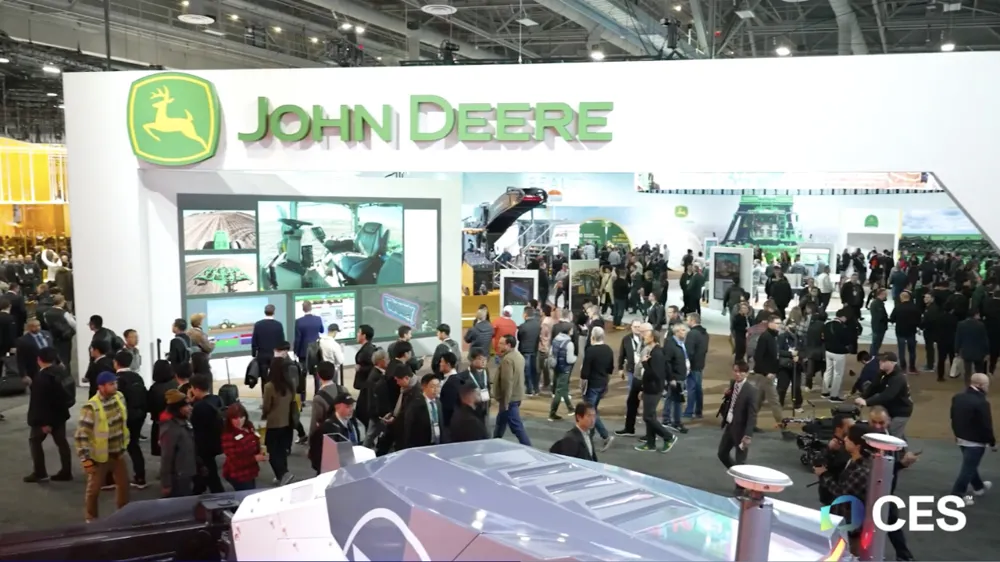 ALT Text: Conference full of people with a big arch with John Deere displayed on top 